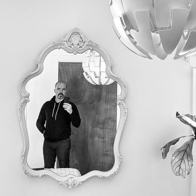 A black and white photo of me taking a picture of myself with my smartphone on a mirror with an ornate frame. A modern pendant lamp is visible in the top right corner, and the edges of a fiddle leaf fig tree near the lower right corner.